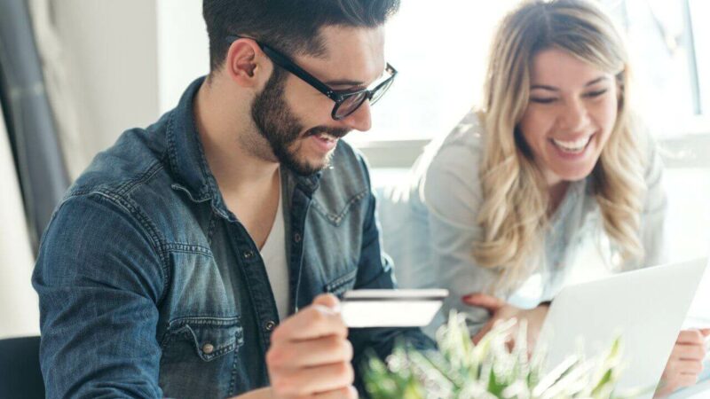 A young couple, smiling while using a laptop, holding a bank card