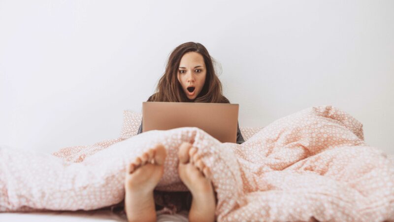 A woman sitting in bed, using a laptop with a surprised look on her face