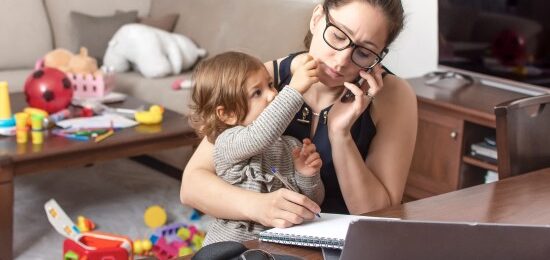 A mother, holding her child while on the telephone and writing notes in a book