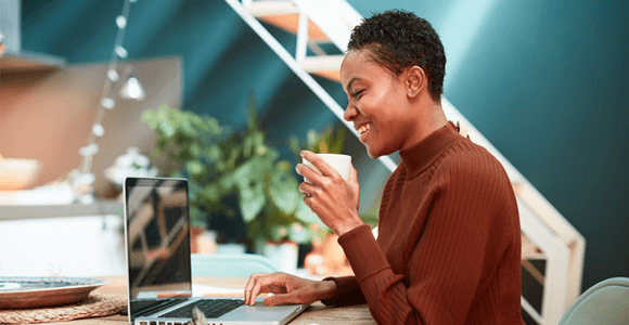 A lady, drinking from a cup and using a laptop